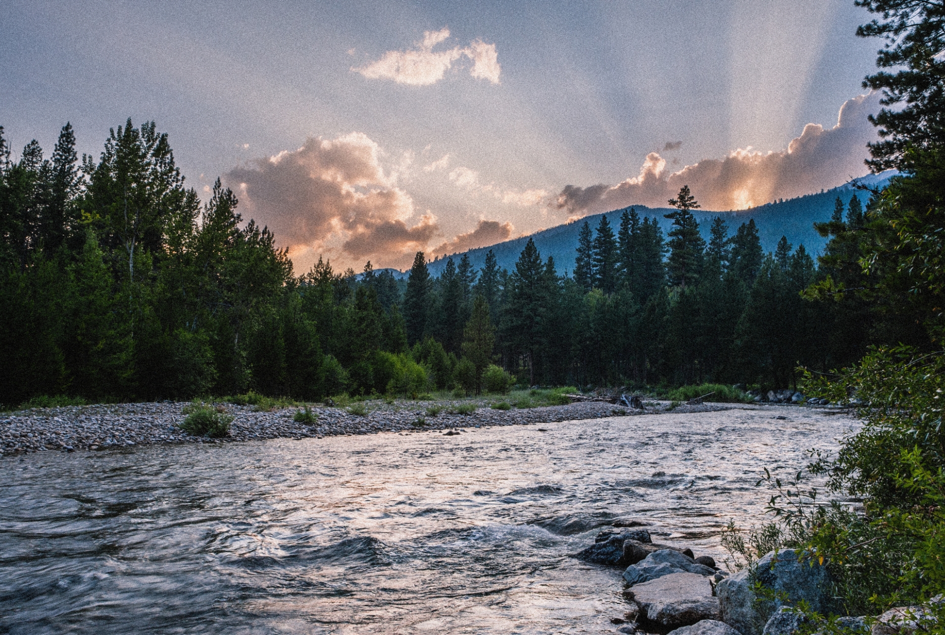 a river at sunset with a mountain in the background and the suns rays peeking through the clouds