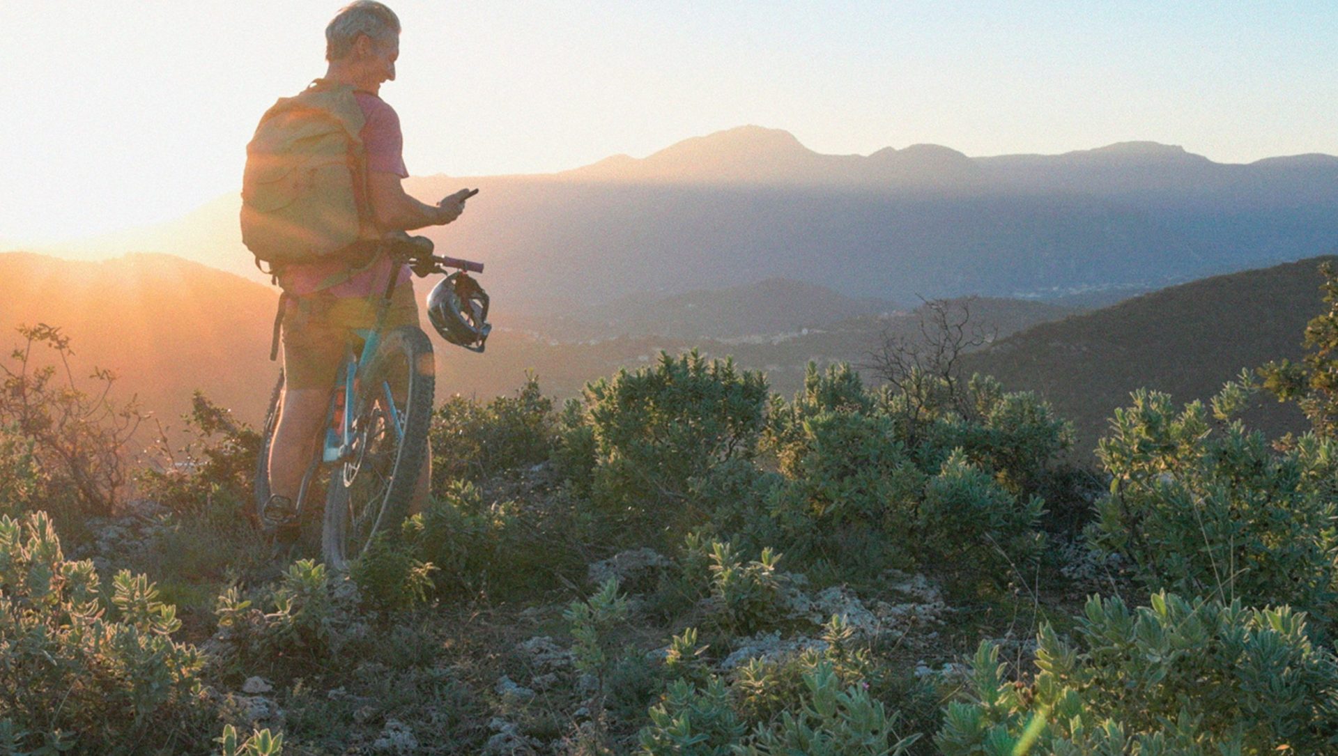 man stops on a cliff ledge to overlook mountain scape during sunrise and look at his phone