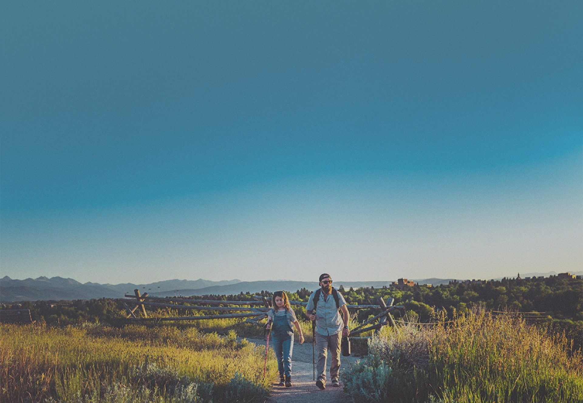 man and woman walking along a trail path with hiking sticks in a grassy plane surrounded by mountains