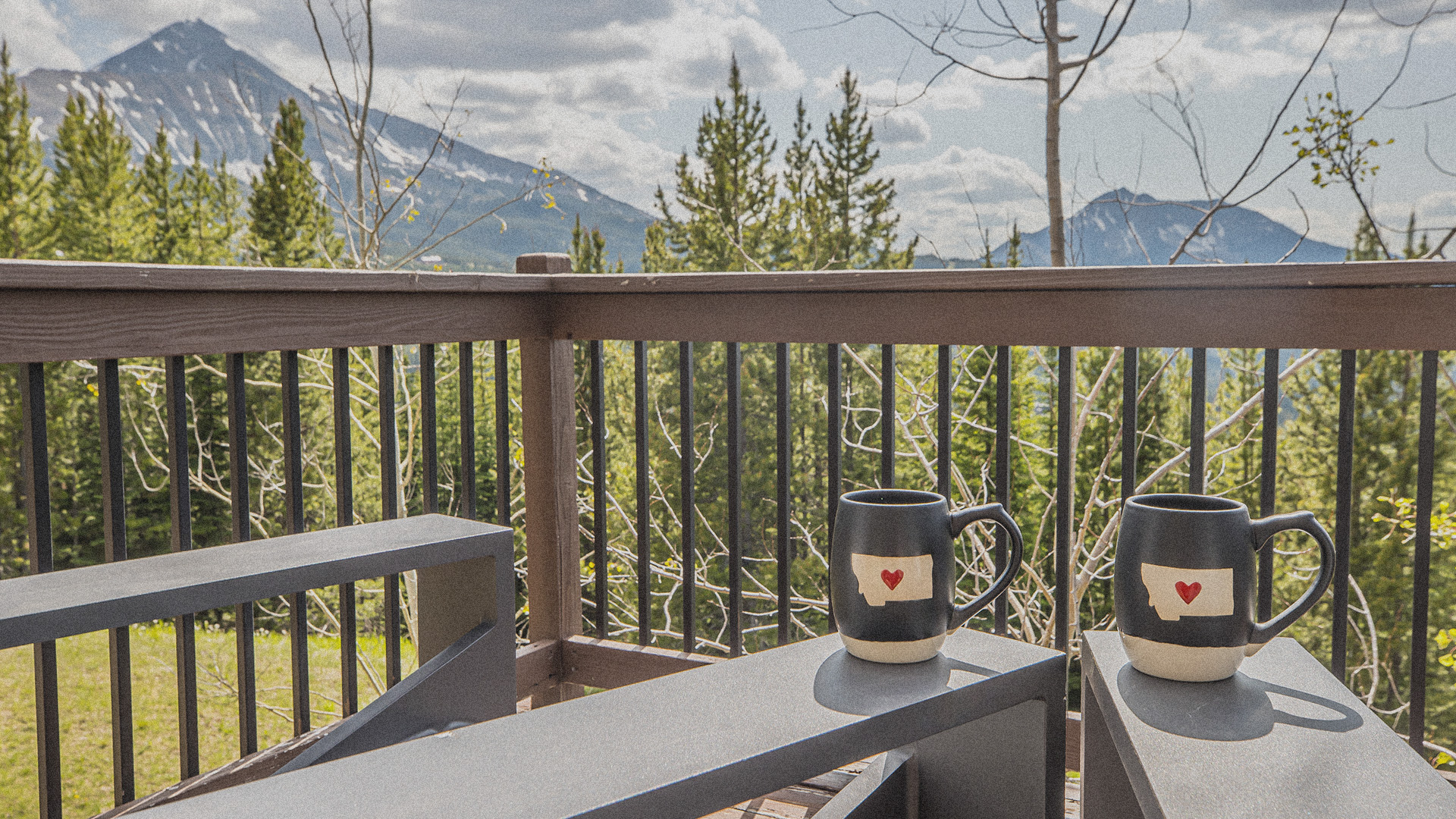 Adirondack chairs on an outdoor patio, facing the mountains, suggest how to live like a local in Big Sky, MT. The mountains are visible in the distance. On the arms of each chair sits a mug with the outline of the state of Montana with a small red heart over it. The background includes lush green trees and grass.
