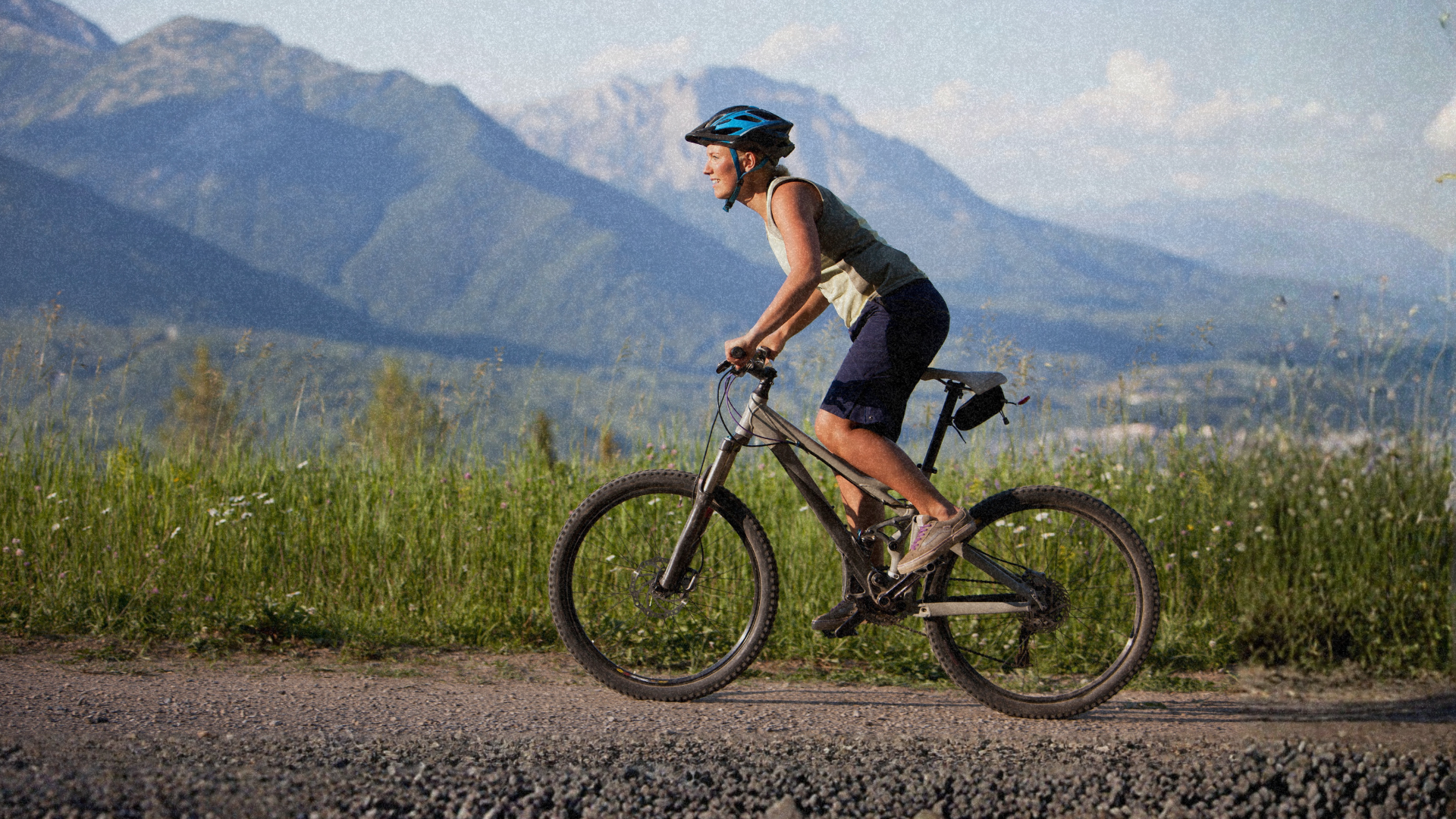A man on a mountain bike wearing sporting apparel and a helmet. He is on an outdoor trail, with the mountains in the background.