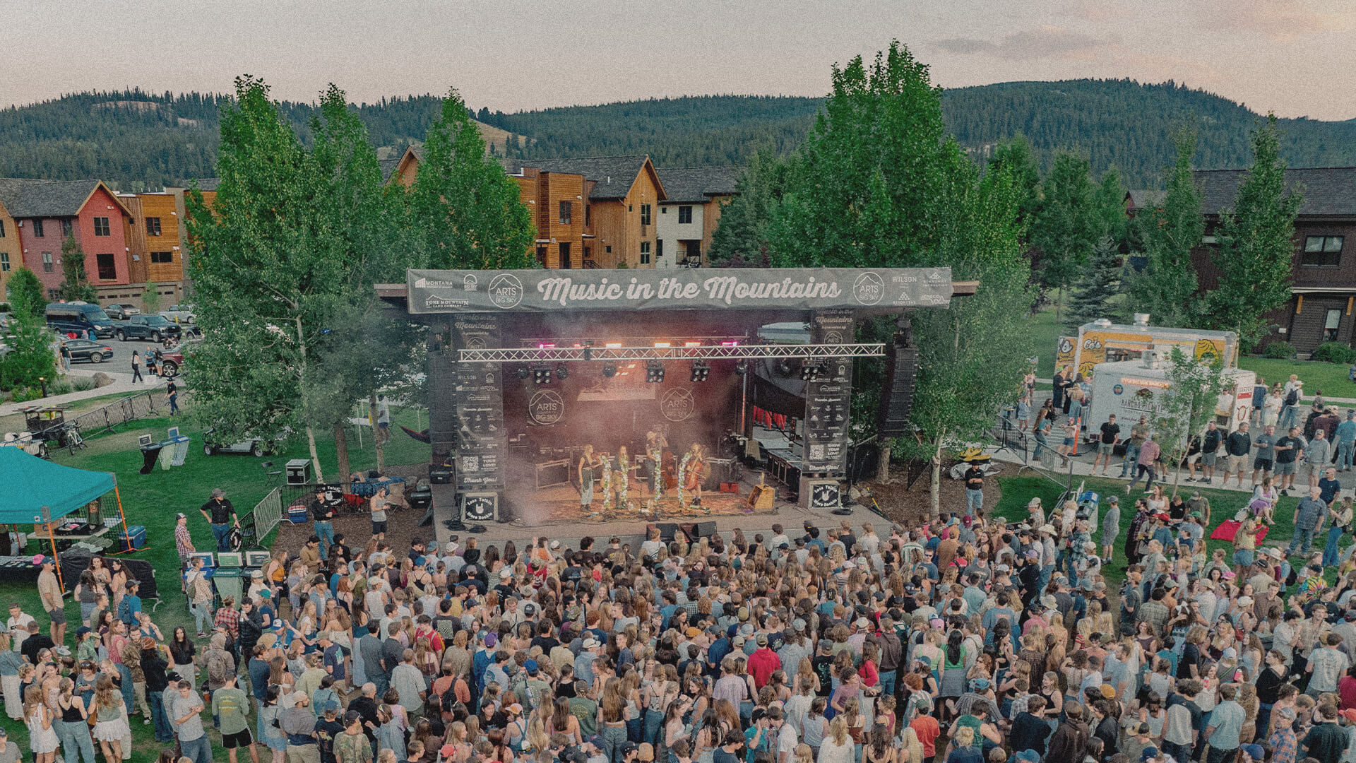 Crowd enjoying a live outdoor concert during the Music in the Mountains summer festival in Big Sky, Montana. The stage is surrounded ny trees and the mountains can be seen in the distance.