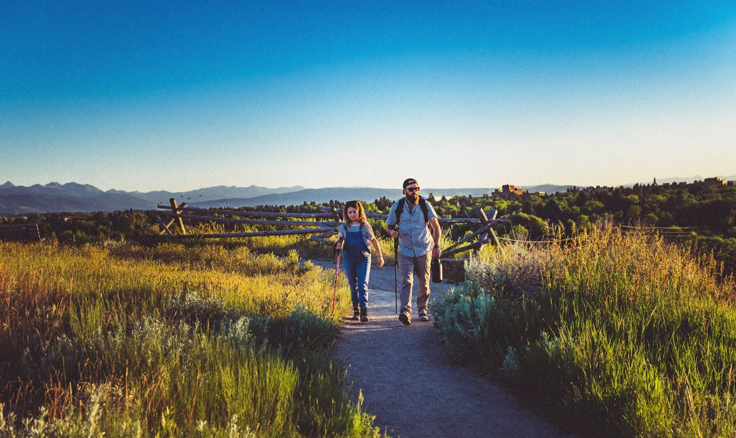 man and woman walking along a trail path with hiking sticks in a grassy plane surrounded by mountains