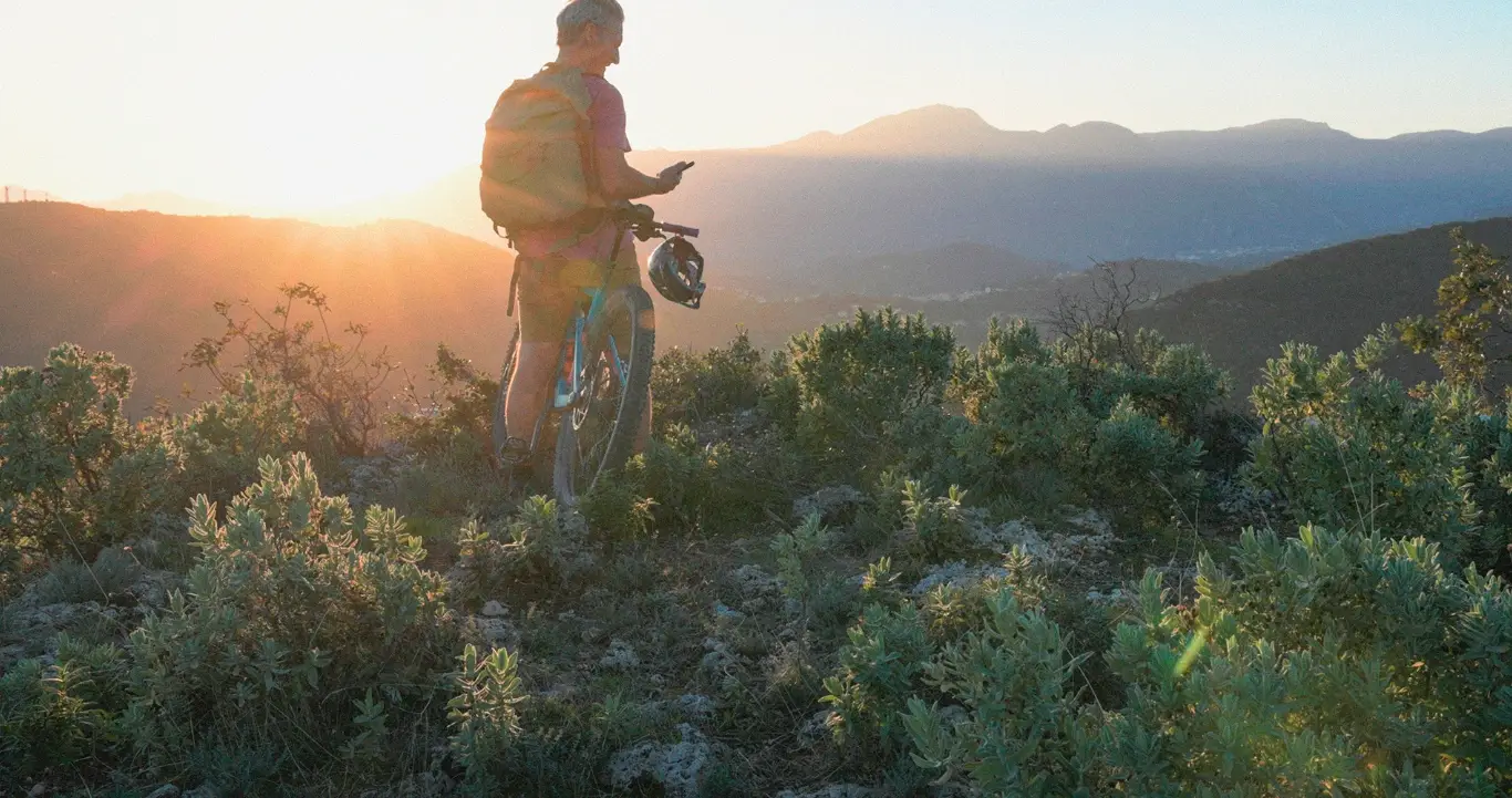 man stops on a cliff ledge to overlook mountain scape during sunrise and look at his phone