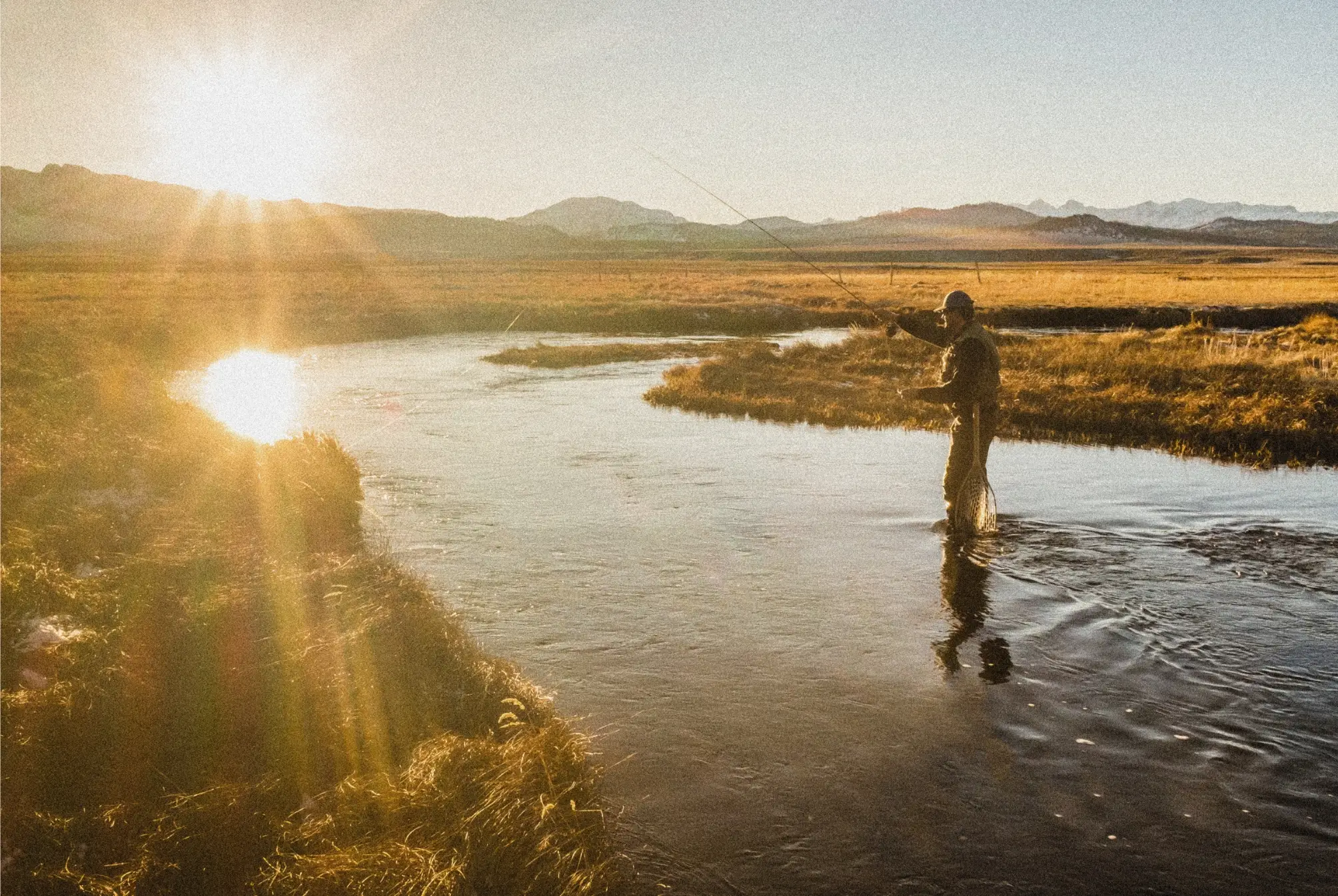 man fishing in a river in front of a mountain range at sun rise