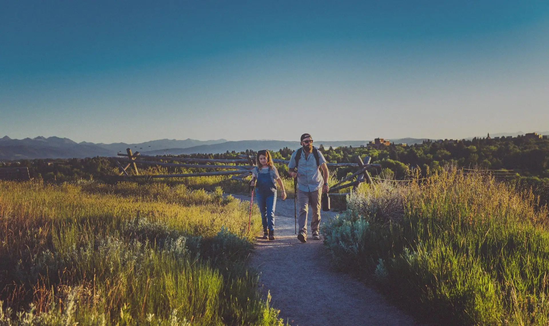 man and woman walking along a trail path with hiking sticks in a grassy plane surrounded by mountains